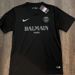 Svart fotbollströja från Nike - Snygg svart fotbollströja från Nike med Balmain Paris-logga och PSG-emblem på bröstet. Tröjan är kortärmad och har en stilren design. Perfekt för fotbollsfans som vill ha något unikt och trendigt.