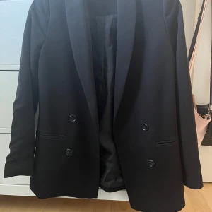 Svart kavaj från Vero Moda - Säljer en stilren svart kavaj från Vero Moda i storlek 34 Kavajen har en klassisk design med dubbelknäppning och två fickor framtill. Perfekt för både vardag och fest!