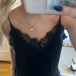 Svart linne med spetsdetaljer - Säljer ett snyggt svart linne med tunna axelband och vacker spetsdetalj vid urringningen. Perfekt för en kväll ute eller en avslappnad dag. Linnet är i ett mjukt och bekvämt material.