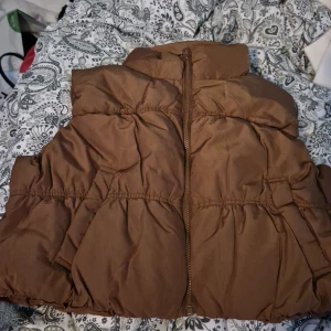 Brun dunväst - Säljer en snygg brun dunväst från Divided. Den har en dragkedja framtill och är perfekt för höst och vinter. Västen är i storlek L och är supermysig att ha på sig när det är lite kyligare ute.