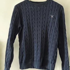 Mörkblå stickad tröja från Gant - Säljer en snygg mörkblå stickad tröja från Gant. Den har ett klassiskt flätmönster och är perfekt för kyliga dagar. Tröjan är långärmad och har ribbade muddar vid ärmslut och nederkant. Perfekt för både vardag och lite finare tillfällen!
