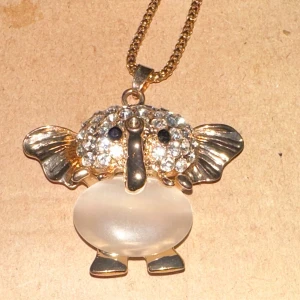 Guldigt elefant-halsband med kristaller - Säljer ett supergulligt halsband med en elefantberlock. Berlocken har en stor, rund kropp i vit sten och huvudet är täckt av glittrande kristaller. Öronen och snabeln är i guldmetall, vilket ger en lyxig känsla. Perfekt för att liva upp vilken outfit som helst!