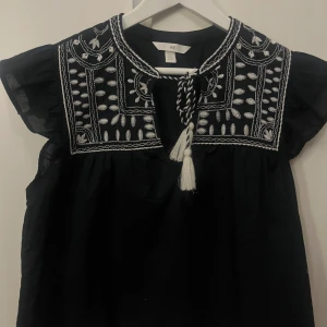 broderad topp - En svart blus från H&M med detaljerad vit brodyr längs bröstet och axlarna. Blusen har korta, luftiga ärmar och knytband framtill med dekorativa tofsar. Perfekt för sommarens varma dagar!