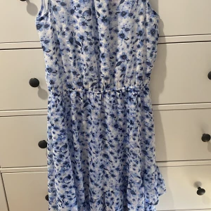 Blommig sommarklänning - Säljer en superfin blommig klänning i ljusblått och vitt. Den är ärmlös och perfekt för sommardagar. Klänningen har en luftig passform med ett elastiskt band i midjan för en bekväm passform. Aldrig använd, lappen är fortfarande på