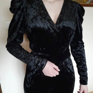 Svart sammet klänning - Säljer en elegant svart jumpsuit. Den har en omlott-design med långa ärmar och en v-ringad hals. Endast använt 1 gång. Perfekt för festliga tillfällen! 