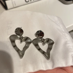 Silvriga hjärtformade örhängen - Säljer ett par snygga silvriga örhängen i form av hjärtan. De har en cool, hamrad yta som ger dem en unik look. Perfekta för att piffa upp vilken outfit som helst, oavsett om det är till vardags eller fest!