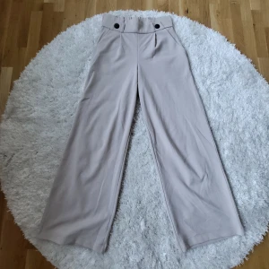 Beige byxor från JDY - Säljer ett par snygga beige byxor från JDY i storlek S. De är högmidjade med en loose passform och har stilrena svarta knappar i midjan. Materialet är en skön blandning av polyester och elastan, vilket gör dem både bekväma och stretchiga. Perfekta för både vardag och fest!