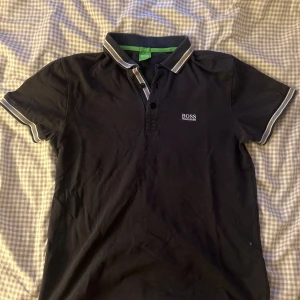 Svart kortärmad pikétröja från Boss - Säljer en stilren svart pikétröja från Boss. Den har korta ärmar och en klassisk krage med vita detaljer. Perfekt för både vardag och fest! 