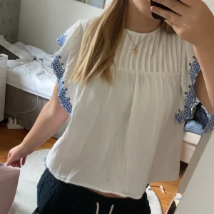 Vit blus med blå broderade detaljer - Säljer en superfin vit blus med korta ärmar och blå broderade detaljer längs kanterna. Blusen har en lös passform och knappar i ryggen. Perfekt för sommardagar eller en avslappnad fest! 🌼