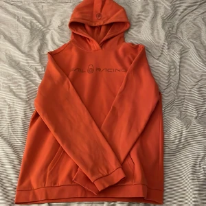 Orange hoodie från Sail Racing - Säljer en cool orange hoodie från Snail Racing. Den har en stor logga på framsidan och en mindre på huvan. Perfekt för höst och vår med sin mjuka och bekväma passform. Den är långärmad och har en klassisk hoodie-stil med ficka framtill.
