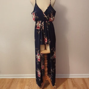 Blommig långklänning med öppen rygg - Säljer en superfin långklänning i mörkblått med blommigt mönster. Klänningen har en v-ringad design och smala axelband som ger en elegant look. Den är perfekt för sommarfester eller en dag på stan. Materialet känns lätt och luftigt, vilket gör den idealisk för varma dagar. Klänningen har också en öppen rygg som ger en extra touch av stil.