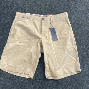 Beige shorts från Tommy Hilfiger - Säljer ett par snygga beige shorts från Tommy Hilfiger i nytt skick. De har en klassisk design med knappar och dragkedja framtill. Tillhörande knappar finns med! Storlek W34  Ny pris är ungefär 900-999kr💕