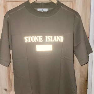 Stone Island Tshirt - T-shirt med reflextryck (se bilderna med/utan blixt) Aldrig haft den på mig, endast packat upp för att ta bilderna men sen skickar jag den så som jag fick den Olivgrön/brun