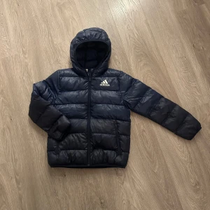 Mörkblå dunjacka från Adidas - Säljer en snygg och varm mörkblå dunjacka från Adidas. Perfekt för kalla dagar med sin quiltade design och praktiska huva. Jackan har en dragkedja framtill och är i mycket bra skick. Passar perfekt för både höst och vinter!
