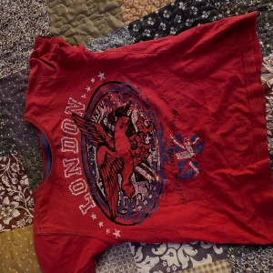 Röd t-shirt med London-tryck - Cool röd t-shirt från KappAhl med ett stort tryck av en örn och texten 'London'. Perfekt för vardagsbruk eller en avslappnad dag ute. T-shirten har korta ärmar och är gjord i mjukt bomullsmaterial. Det är egentligen en pojktröja i storlek 128 men passar för xs/s