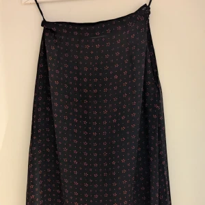 Svart kjol med små blommor från Cappuccini - Säljer en tunn svart långkjol från Cappuccini i storlek M. Längden är ca 79 cm. Mått i midjan ca 69 cm. Knäpps med liten knapp och dragkedja i sidan. 32 cm slitsi båda sidorna. Består av två lager där det yttre lagret är i mesh. Kjolen har ett sött blommönster i rött och är tillverkad i 100% polyester. Perfekt för både vardag och festliga tillfällen. Den är i mycket bra skick och redo för en ny garderob! 🌸