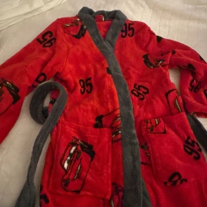 Cars morgonrock för barn - Röd med Blixten McQueen-motiv - Storlek 110/116 - En mysig och varm morgonrock för barn i röd färg med gråa detaljer. Designen är inspirerad av Disney Cars, med Blixten McQueen-tryck och nummer 95 på tyget, perfekt för små fans av filmen. nödtan nytt💫