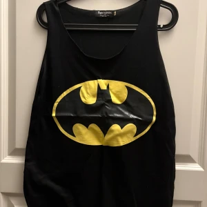Svart linne med Batman-logga från Fancyqube - Säljer ett svart linne från Fancyqube med den ikoniska Batman-loggan i gult på framsidan. Perfekt för alla äventyr eller som en cool detalj till vardagsoutfiten. Linnet är i bra skick och redo för en ny ägare! Kommer från djur och rökfritt hem 