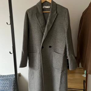 Grå kappa från Zara - Säljer en stilren grå kappa från Zara i storlek L. Den är i mycket bra skick och perfekt för höst och vinter. Kappan har en klassisk design med långa ärmar och två fickor framtill. Den är gjord i ett ullblandat material som håller dig varm och snygg. Perfekt för både vardag och fest!