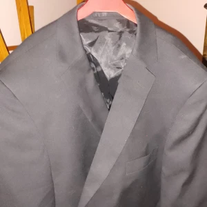 Kavaj ifrån Dressmann - Snygg svart kavaj i klassisk stil. Perfekt för både formella och semi-formella tillfällen. Kavajen har en enkel knäppning och är i ett material som ser ut att vara av hög kvalitet. Passar bra till både byxor och jeans för en stilren look.