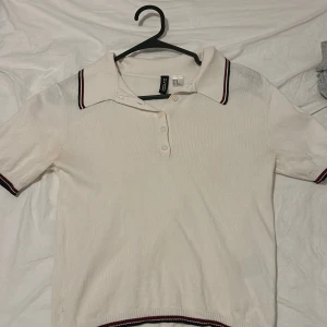 Vit pikétröja från Divided - Säljer en stilren vit pikétröja från Divided i storlek XS. Tröjan har korta ärmar och en klassisk krage med röd och svart randdetalj. Perfekt för en casual look eller en dag på stan. Knappar framtill för en snygg finish. 🖤