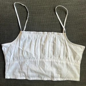 Vit ärmlös linnetopp - Säljer en söt vit linnetopp med smala axelband. Toppen har en rynkad design framtill och smockdetaljer på baksidan för en bekväm passform. Perfekt för sommardagar eller som ett lager under en kofta. Passar till både jeans och kjol! Står 42 men är som en S-M