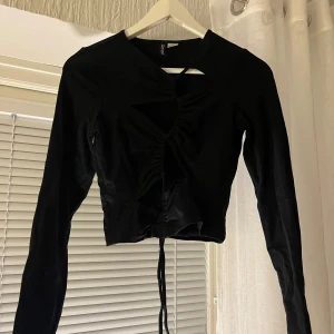 Svart långärmad topp från Divided - Snygg svart långärmad topp från Divided med knytning framtill. Den har en unik design med rynkade detaljer och en liten öppning vid halsen. Perfekt för en kväll ute eller en fest. Toppen är i bra skick och passar till många olika outfits.