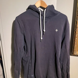 Mörkblå Morris Hoodie - Säljer en snygg mörkblå hoodie med en liten broderad detalj på bröstet. Den har en justerbar huva med vita snören och långa ärmar. Perfekt för en avslappnad stil under höst och vinter. Passar både till vardags och chillkvällar.