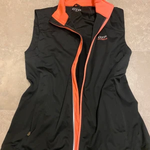 Svart och orange väst från OTP Extreme - Säljer en svart väst från OTP Extreme med orange detaljer och dragkedja. Västen har en hög krage och är perfekt för träning eller utomhusaktiviteter. Den är i mycket bra skick och har praktiska fickor med dragkedja. Perfekt för höst och vår!
