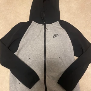 Grå och svart hoodie från Nike - Säljer en snygg och bekväm grå och svart hoodie från Nike. Den har en dragkedja framtill och en klassisk Nike-logga på bröstet. Perfekt för både träning och vardag. Huvan är justerbar och tröjan har praktiska fickor. Passar perfekt till höst och vår!