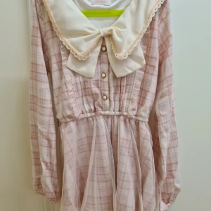 Rutig rosa tröja med rosett Liz Lisa - Söt rosa tröja med rutigt mönster och en stor vit rosett vid halsen. Tröjan har långa ärmar och knappar framtill. Flanell liknande tyg, perfekt för kallare väder. One size, skulle säga S. Helt ny, endast provad. 