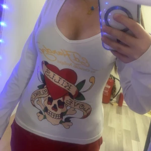 Vit långärmad Edhardy topp - Säljer en vit långärmad topp med ett coolt tryck av en dödskalle och hjärta med texten 'Love Kills'. Perfekt för en avslappnad stil med en edgy touch. Toppen är i bra skick och passar perfekt till jeans eller kjol.