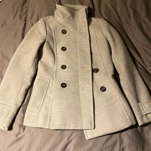 Grå kappa med knappar - Säljer en jättefin grå kappa i mycket bra skick! Den har en dubbelknäppt design med stora svarta knappar och en hög krage. Perfekt för höst och vår.