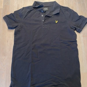 Svart pikétröja från Lyle & Scott - Säljer en svart pikétröja från Lyle & Scott i storlek 12/13 år. Tröjan har en klassisk krage med knappar och ett broderat gult emblem på bröstet. Perfekt för både vardag och lite finare tillfällen. Den är i bra skick och redo för en ny ägare!