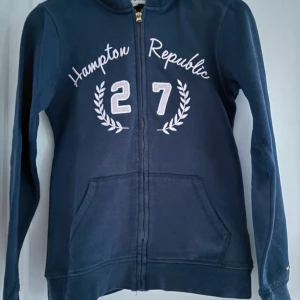 Mörkblå hoodie från Hampton Republic - Säljer en snygg mörkblå hoodie från Hampton Republic med dragkedja och tryck framtill. Den är i storlek 158 och gjord av 80% bomull och 20% polyester, vilket gör den både bekväm och hållbar. Perfekt för höst och vår! Köparen betalar frakt. 