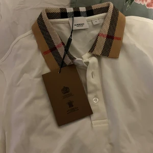Vit pikétröja från Burberry - Säljer en snygg vit pikétröja från Burberry med den klassiska rutiga kragen i beige, svart och röd. Tröjan är i nytt skick och perfekt för både vardag och fest. Den har en normal passform och är tillverkad i bomull för en skön känsla. Passa på att köpa denna stilrena tröja!