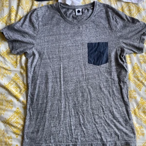 Grå t-shirt med ficka från NN07 - Säljer en stilren grå t-shirt från NN07 i mycket bra skick. Den har en mörkare grå bröstficka som ger en snygg kontrast. Perfekt för vardagsbruk eller en avslappnad dag ute. Passar till det mesta och är superbekväm!