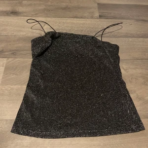 Svart glittrig tubtopp - Säljer en snygg svart glittrig tubtopp med tunna axelband. Perfekt för festliga tillfällen! Toppen har en enkel och stilren design som gör den lätt att matcha med olika outfits. Den är i bra skick och redo för nästa ägare.