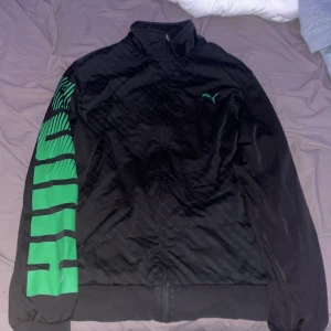 Svart träningsjacka från Puma - Säljer en svart träningsjacka från Puma i bra skick. Den har en dragkedja framtill och långa ärmar med gröna detaljer och text på ena ärmen. Perfekt för träning eller som en sportig vardagsjacka. Stor logga på bröstet i grönt.
