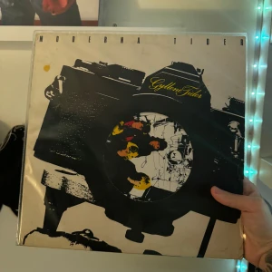Moderna Tider av Gyllene Tider - Säljer ett klassiskt vinylalbum 'Moderna Tider' av Gyllene Tider. Omslaget har en cool design med en stor kamera och bandets namn i gult. Perfekt för samlare eller fans av svensk popmusik!