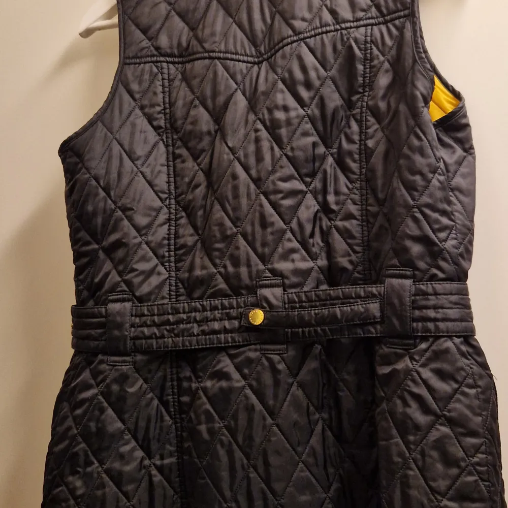 Säljer en snygg svart quiltad väst från Barbour i storlek EUR 34. Västen har gul insida och detaljer som knappar och bälte i midjan. Perfekt för höst och vår, med en stilren look som passar både till vardags och fest.. Jackor.