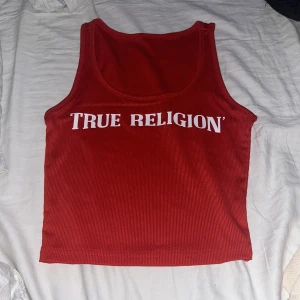 Röd ribbad linnetopp från True Religion - Säljer en snygg röd ribbad linnetopp med trycket  True Religion. Den är ärmlös och har en djup rund halsringning. Perfekt för sommardagar eller en avslappnad festkväll. Toppen är croppad och har en cool, casual stil. Passar perfekt med jeans eller shorts!