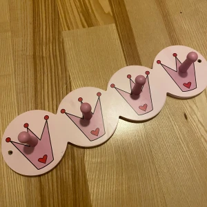 Rosa väggkrok med kronmotiv - Söt väggkrok i rosa med fyra krokar, perfekt för barnrummet. Varje krok är dekorerad med en krona och ett litet hjärta, vilket ger en prinsessliknande känsla. Tillverkad i trä och lätt att montera på väggen.