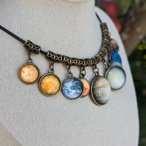 Halsband med planetmotiv - Unikt halsband med hängande berlocker som föreställer olika planeter i solsystemet. Berlockerna har en antik bronsfärgad ram och hänger på en svart rem. Perfekt för rymdälskare.