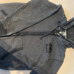 Grå hoodie från Roots - En snygg och bekväm grå hoodie från Roots. Den har en dragkedja framtill och en justerbar huva. Perfekt för kalla dagar och har en broderad logga på bröstet. Supermysig och stilren! Populärt kanadensiskt märke som inte går att köpa i sverige.