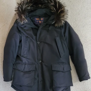 Woolrich dunjacka, Vinterjacka, Arctic Parka - Vinterjacka i dun från Woolrich i mycket gott skick. Fyra rymliga fickor på framsidan, två utanpåliggande och två tvärfickor, samt två stora innerfickor. Tvåvägsdragkedja, dold dragsko i midjan, avtagbar pälskant på luvan, dold mudd i ärmslut.  