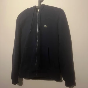 Zip hoodie från Lacoste  - Säljer en stilren marinblå hoodie från Lacoste. Den har en dragkedja framtill. Perfekt för höst och vinter med sin mysiga passform och långa ärmar. Ett måste för garderoben!