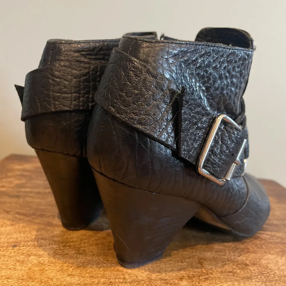 Snygga svarta boots i skinn med en cool spetsig tå och klack. De har en bred rem med ett stort metallspänne som ger en edgy look. Perfekta för höst/vår, och de passar både till vardags och fest. Skorna har en dragkedja på sidan. Fin ny sula.. Kengät.
