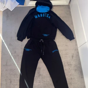 Svart och blå hoodie från MAN - Säljer en snygg svart Tracksuit från bohooMAN med blå detaljer. Den har en stor, bekväm huva med blå halsvärmare i insidan och matchande blå text på bröstet. Perfekt för en avslappnad stil eller träning. Passar bra till höst och vinter! Strlk M