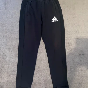 Svarta mjukisbyxor från Adidas - Säljer ett par svarta mjukisbyxor från Adidas. De är superbekväma och perfekta för träning eller chill. Byxorna har elastisk midja och två fickor med dragkedja. Adidas-loggan syns tydligt på ena benet. Gjorda av Primegreen-material, vilket är ett hållbart val!